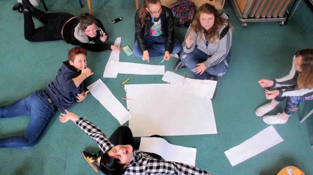 young people and volunteers sitting in a circle on the floor smiling and giving thumbs up to the camera above them
