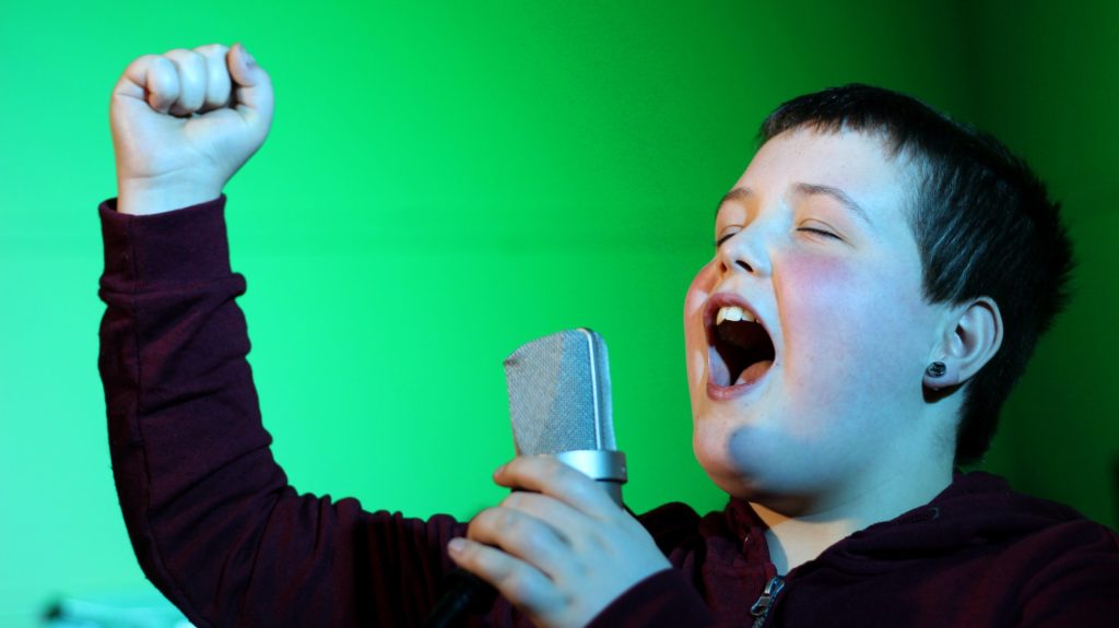 Young boy holding microphone up to his wide open mouth singing, eyes closed, holding his fist in the air dramatically.
