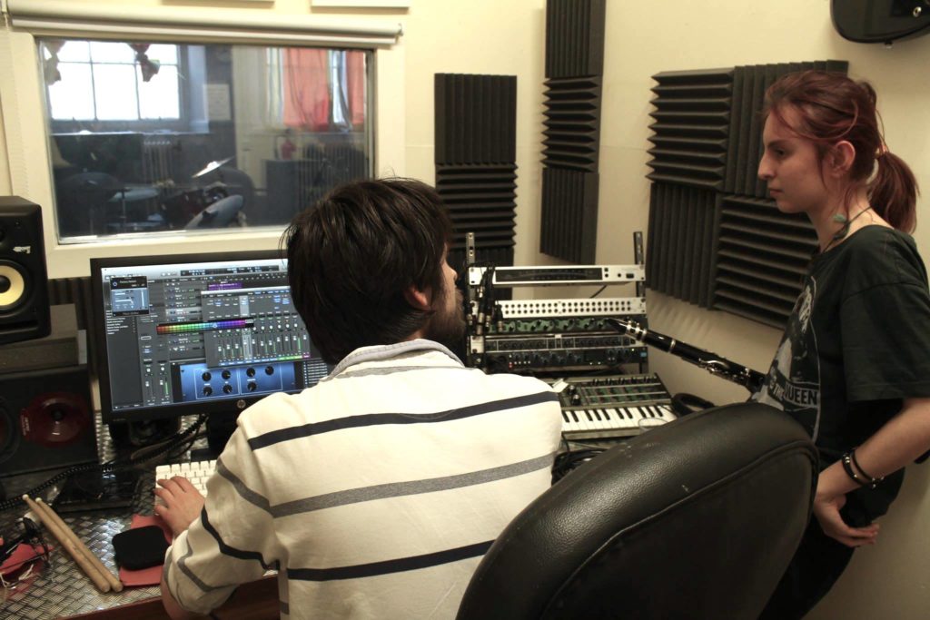Tutor and young person in recording studio with equipment, soundproofing and a window leading to recording room