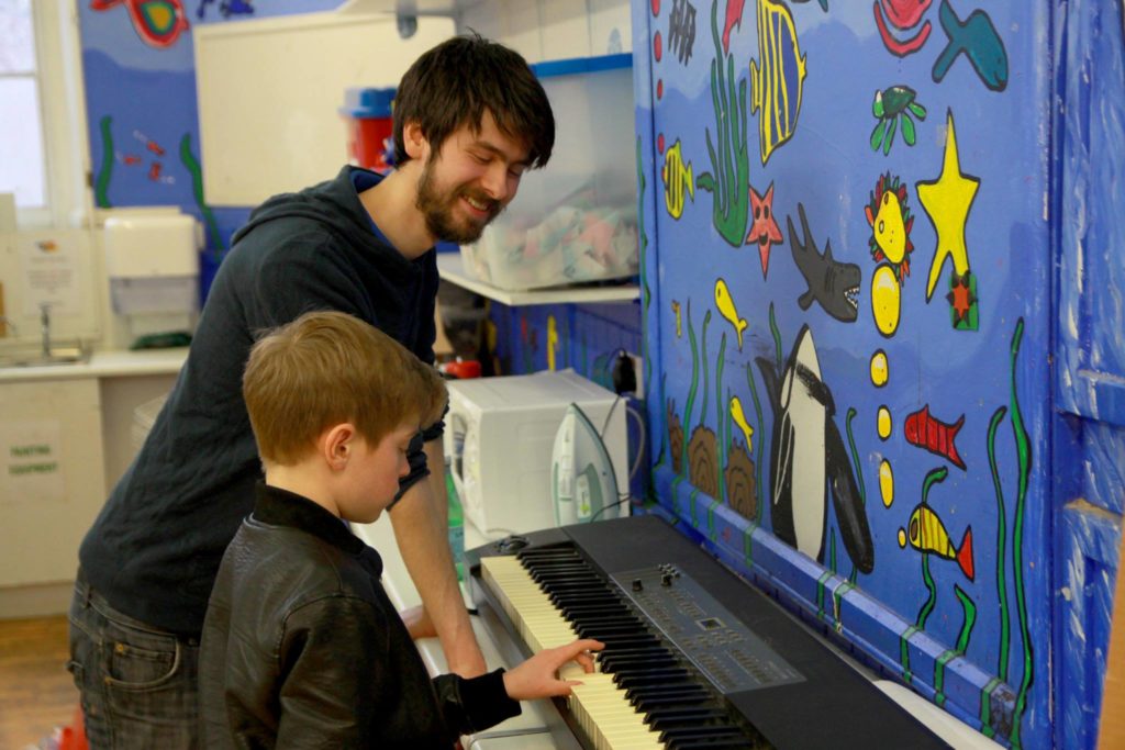 Young person concentrating and playing the keyboard, while tutor watches on, smiling